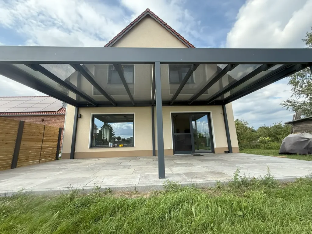 Ein wunderschön gestalteter Garten an einem sonnigen Sommertag. Im Vordergrund eine moderne, anthrazitfarbene Terrassenüberdachung aus Stahl mit stilvollen Gartenmöbeln im Schatten. Im Hintergrund gepflegter Rasen und große, schattenspendende Bäume. Die Atmosphäre ist entspannend und einladend. Fotorealistisch, 8k.