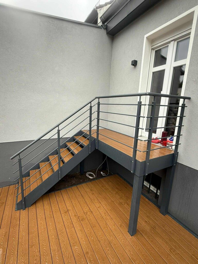 Montageprozess: Ein Montageteam in Arbeitskleidung hebt gerade ein großes Stahlmodul mit einem kleinen Kran an seinen Platz. Man sieht die präzise Vorfertigung der Teile. aussentreppe AUS WPC DIELEN